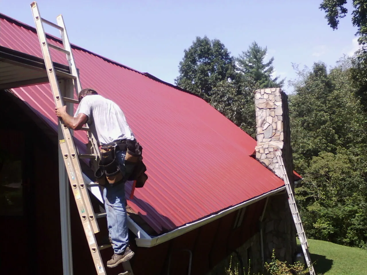 Roofer on a ladder beside a red metal roof and stone chimney — Soffit & Fascia Repair by Roofing Brothers in Red Wing